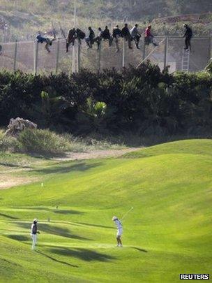 A golfer hits a tee shot as African migrants sit atop a border fence, during an attempt to cross into the Spanish North African enclave of Melilla