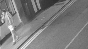 A black and white CCTV image, showing a person walking on a pavement.