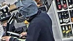 A masked man is pointing a knife towards a member of staff behind the counter. He is holding a green carrier bag.