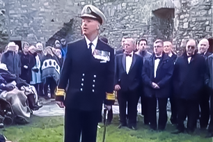 Dyn mewn gwisg llyngesydd yn sefyll ar wair gyda chynulleidfa o'i gwmpas. Mae'n gwisgo het wen, siaced a throwsus dy, crys gwyn a thei du. Mae ganddo sawl medal ar ei siaced a chleddyf yn ei law chwith