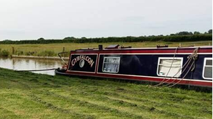 A narrowboat on the Grand Union Canal with green grass in front and on the other side of the water. The boat is blue and purple with its name painted around a window