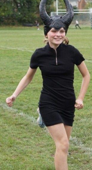 A girl wears black running clothes and black horns on her head. She is running on a grass track.
