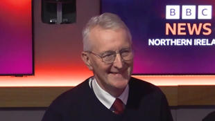 Hilary Benn sitting in Radio Ulster studio booth in front of a microphone. He is wearing a dark pullover, white shirt and red tie. He has round glasses and short grey hair. Behind him are two screens with the BBC News Northern Ireland logo on them on purple/red backgrounds.