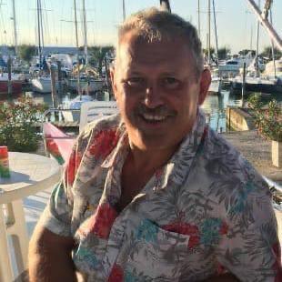 Mike O'Leary wears a tropical shirt, smiling in the sunshine at a harbour, boats visible in the background