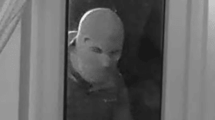 A black and white image of a masked person peering through the glass door. The person is holding something in their right hand.