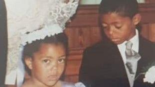 A photo of Leroy and his sister as young children. He is wearing a black suit with a white shirt and white flower pinned to his suit jacket. She is wearing a white headband with a ribbon at the back and a purple dress. Leroy is looking down at Natalie, who is looking at the camera. 