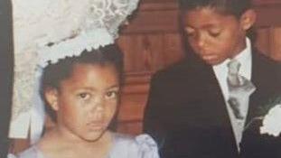 A photo of Leroy and his sister as young children. He is wearing a black suit with a white shirt and white flower pinned to his suit jacket. She is wearing a white headband with a ribbon at the back and a purple dress. Leroy is looking down at Natalie, who is looking at the camera.
