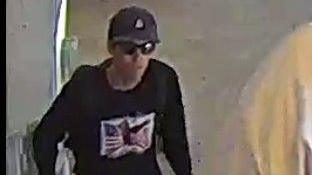 Image shows a slim, white man wearing sunglasses, a dark baseball cap, and a dark sweater with a picture of an American flag on the front.  
