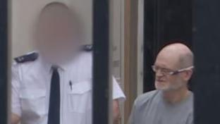 Two men are in the picture. The one on the right is John Garrett O'Sullivan. HE is baldin with a grey beard and glasses. He is wearing a grey, round neck top. He is beside a man who has his face blurred and is wearing a white shirt and tie.