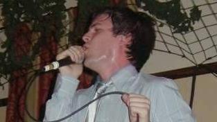 Brandon Flowers passionately singing into a microphone. He is wearing a white shirt and silver tie and holding the microphone in one hand and its cord in the other. The side of his face is coated with sweat.