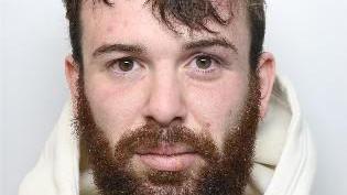 A mugshot of a bearded man who is wearing a white hoodie