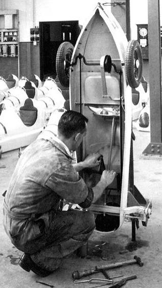 An example of the adaptive technology used to help the ex-miners. A jig was used to hold the pedal cars in place vertically while workers assembled them. The image is a black and white photo of a man crouching to work on the car supported by the rig.
