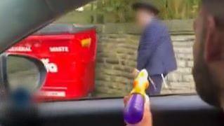 Still of a man in a car spraying a water pistol at a boy walking past who wears an Orthodox Jewish hat. 