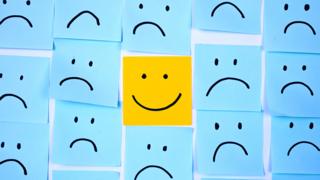 A wall covered in blue post it notes with sad faces on them. In the centre, sits a yellow post-it note with a smiley face.