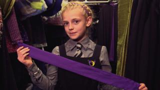 A girl wearing a school uniform holding a purple sash in front of her surrounded by costumes, Sybil from the Worst Witch.