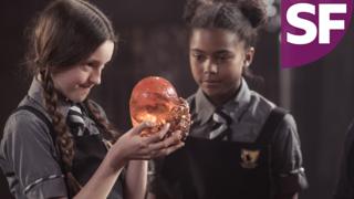 A girl holding a spherical orange stone while another girl looks at it, Mildred Hubble and the Founding Stone.