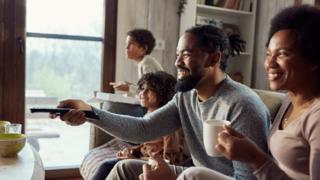 A family sitting down to watch TV, with a man holding a TV remote and woman holding a mug, the children are in the background