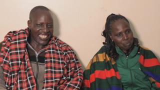 Simion beams while wearing a plaid jacket over a zip up top and his wife, wearing a green jacket has tears in her eyes