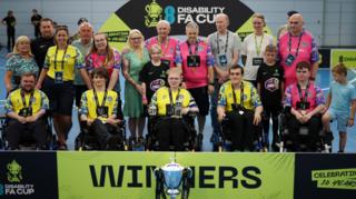 Team shot of Leeds PFC with the trophy standing before them. A yellow and black banner is behind the trophy which reads WINNERS. The players sit in their wheelchairs, while coaches, family and friends stand behind.