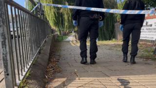 The bottom half of two officers in dark clothes walking beyond blue and white police tape on a footpath.