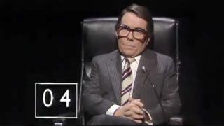 Ronnie Corbett wearing a suit and sitting in the Mastermind chair. There's a small score counter in the bottom left corner