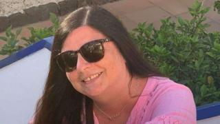 Lesley McTear has long dark hair and wears sunglasses and a pink top. She is smiling and appears to be outside.