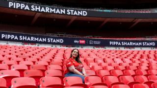 A picture of Pippa Sharman, wearing a red Every Minute Matters T-shirt, sat in Wembley stadium with "Pippa Sharman Stand" behind her on screens running behind her.