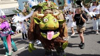 A green and brown carnival fantasy creature with horns and yellow eyes sticks an oversized pink tongue out at the camera. It is a sunny day with lots of other people dressed up for the carnival procession.