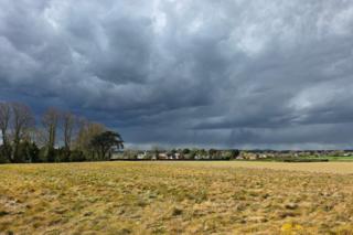 Dark storm clouds over a rural landscape, with rain falling in the distance beyond a grassy field and a line of trees.