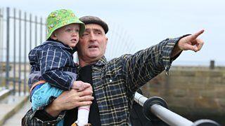 A man and a young boy by the seaside, the man is pointing something out.