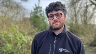 Jack Cripps is wearing glasses and a black zip-up fleece with the Suffolk Wildlife Trust logo. He is standing in a woodland and looking into the camera.