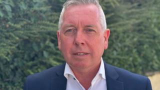 A man with short grey hair, a navy jacket and a white shirt.