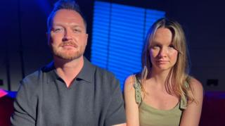 A man and a woman look at the camera. He has a ginger beard and is wearing a grey short-sleeved top. She has long blonde hair and is wearing a green top