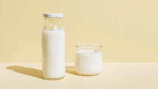 A glass bottle of milk and small glass of milk next to it. With a pale yellow background.