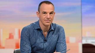 Martin Lewis sat in a TV studio. He's looking at the camera and wearing a dark blue shirt.