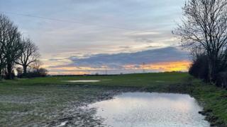 The sun sets in a pale blue sky over a flooded field in the foreground