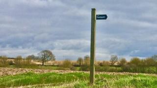 A cloudy sky behind a green field with a footpath sign in the centre.