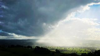 Half the sky over a view of fields is filled with dark blue-grey clouds while the sun is breaking out behind and filling the other half with a white glow