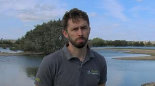 Jamie Parsons poses for the camera with a glum expression. A wetlands with a very low water level is visible behind him.