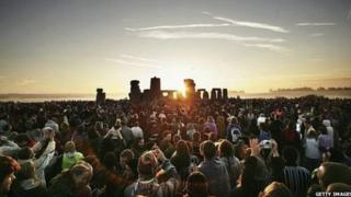 A large crowd are standing in front of Stonehenge as the sun rises on the summer solstice