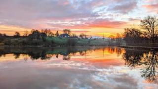 The sun is rising over the horizon with pink and purple clouds above. It's all reflected in the water below which is edged with trees