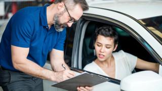 A young woman with short black hair and a white top sits in the driver's seat of a white hatchback, and looks at paperwork on a clipboard held by the car salesman, who wears a blue t-shirt and glasses. 