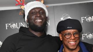 Stormzy wears a black hoodie and a festive hat with antlers. He has his arm around Ian Wright, who is wearing black glasses and a black cap.