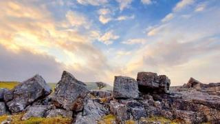 A view of a golden and blue sky with clouds over a natural rock wall with a field behind it.