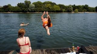 A man backflips into a river from a pontoon, wearing swim shorts. A woman sits on the pontoon with her back to the camera, wearing a bikini