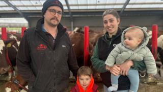 Farmers Adam Johnstone, wearing a black gilet and black hat and Lucy Johnstone, wearing a green fleece and black gilet, standing in front of a cow shed, with Lucy holding her 5-month-old son and their 4-year-old son standing in between them