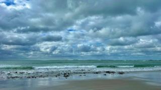 Blue and grey full clouds fill the sky above a greenish sea with sand and some rocks on the foreshore