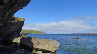 Craggy rocks frame the left side of the picture with blue sea, mountains in the background and blue sky.