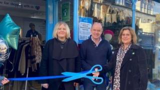 Two women and a man stand outside a new charity shop, smiling as they cut a ribbon with big blue scissors