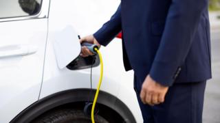 A person, whose face is out of frame, is charging a white electrical vehicle. They are wearing a navy suit.